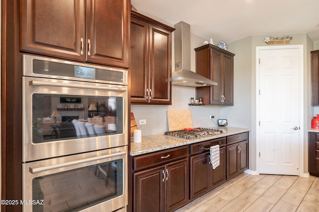 kitchen with wall chimney range hood, appliances with stainless steel finishes, dark brown cabinets, and light stone countertops