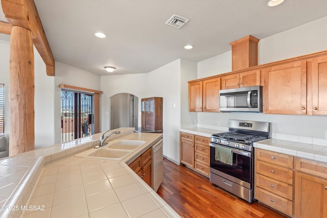 kitchen featuring appliances with stainless steel finishes, a sink, arched walkways, tile counters, and dark wood finished floors