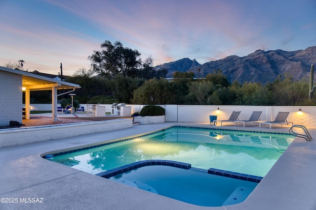 pool at dusk featuring a pool with connected hot tub, a patio area, fence, and a mountain view