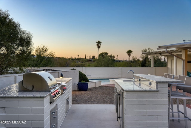 patio terrace at dusk with a fenced backyard, area for grilling, outdoor wet bar, and a sink