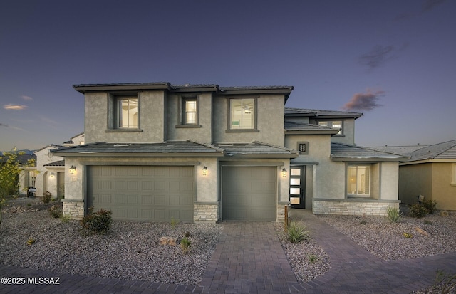 prairie-style home featuring an attached garage, decorative driveway, stucco siding, and stone siding