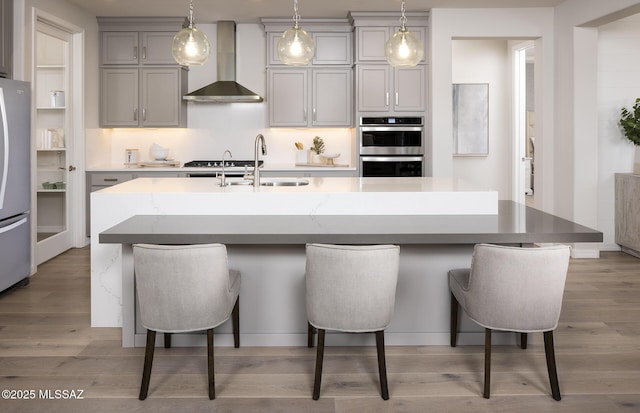 kitchen with wall chimney range hood, gray cabinetry, a sink, stainless steel appliances, and light wood-style flooring