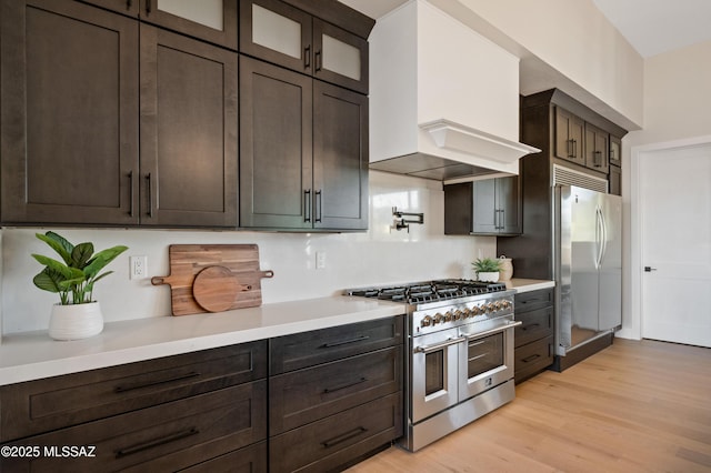 kitchen with appliances with stainless steel finishes, premium range hood, light countertops, light wood-style floors, and dark brown cabinets