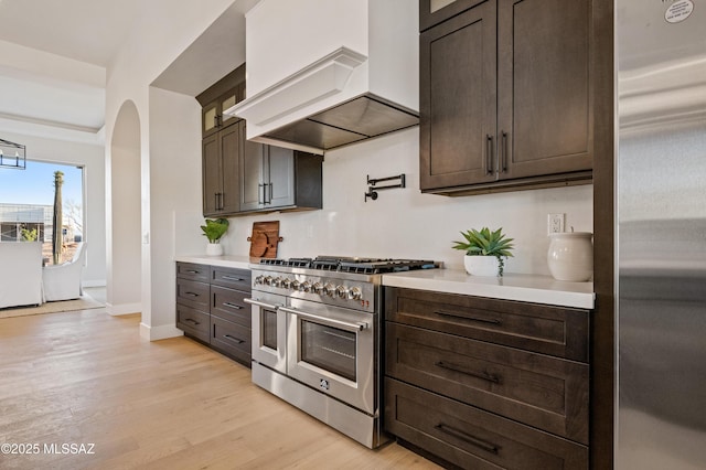 kitchen featuring appliances with stainless steel finishes, premium range hood, light countertops, light wood-type flooring, and dark brown cabinets