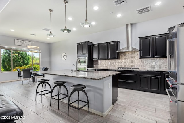 11874 N Raphael Way, Tucson, AZ 85742 - kitchen featuring wall chimney range hood, dark cabinets, appliances with stainless steel finishes, decorative backsplash, and light stone counters