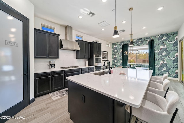 1144 S Castar Dr, Tucson, AZ 85745 - kitchen featuring wall chimney exhaust hood, stainless steel gas stovetop, an island with sink, light wood-style floors, and dark cabinetry