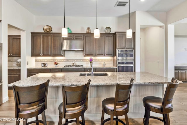 31618 S Tamarisk Pl, Oracle, AZ 85623 - kitchen featuring under cabinet range hood, appliances with stainless steel finishes, tasteful backsplash, dark brown cabinetry, and a kitchen island with sink