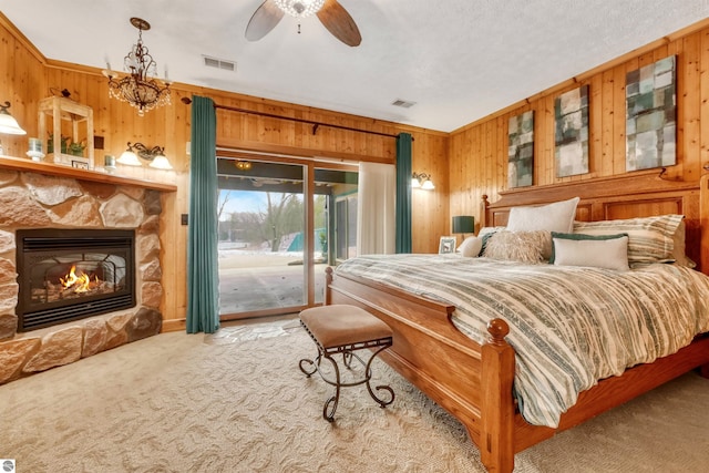 carpeted bedroom with a fireplace, wooden walls, ceiling fan with notable chandelier, and access to exterior