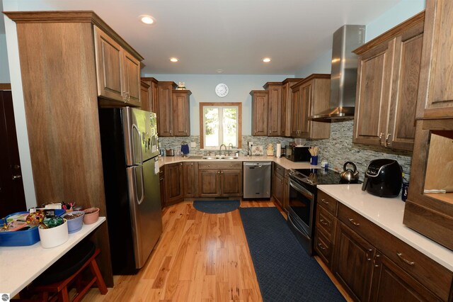 kitchen with appliances with stainless steel finishes, wall chimney range hood, sink, decorative backsplash, and light wood-type flooring