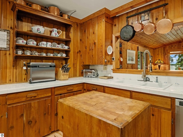 kitchen featuring tile counters, sink, stainless steel dishwasher, wooden walls, and a kitchen island