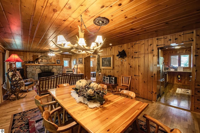 dining room with ceiling fan with notable chandelier, wooden ceiling, hardwood / wood-style floors, wooden walls, and a stone fireplace