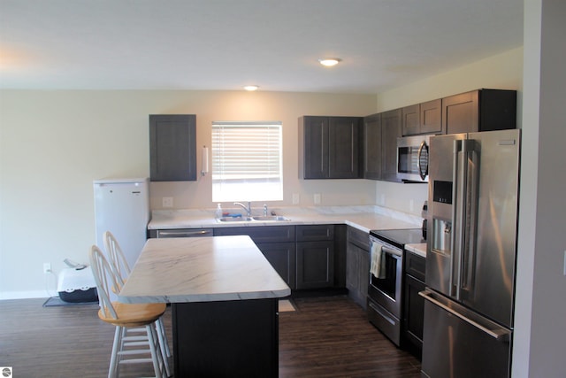 kitchen with appliances with stainless steel finishes, light countertops, a sink, a kitchen island, and dark wood-type flooring