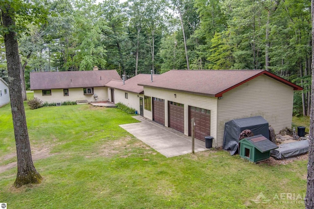 single story home with a garage, a shingled roof, driveway, a front lawn, and a wooded view