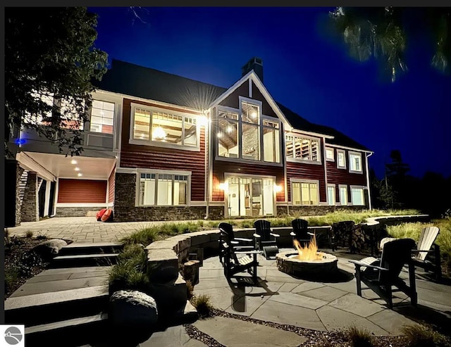 11305 Evergreen, Charlevoix, MI 49720 - back of house at twilight featuring a fire pit, a chimney, stone siding, and a patio
