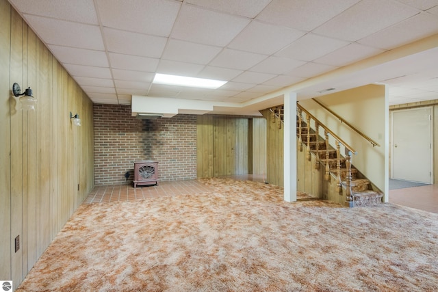 basement with light carpet, a paneled ceiling, wood walls, and a wood stove