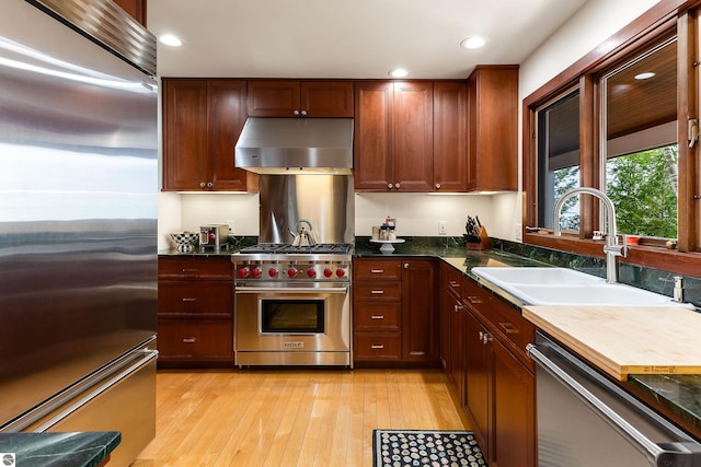 kitchen with sink, exhaust hood, dark stone countertops, high quality appliances, and light hardwood / wood-style floors