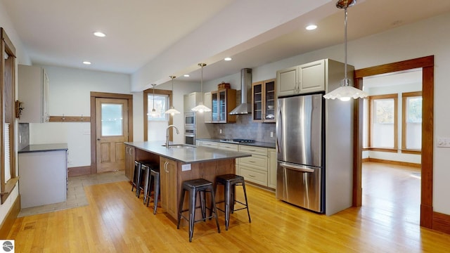 kitchen with wall chimney range hood, light hardwood / wood-style flooring, a center island with sink, a kitchen bar, and pendant lighting