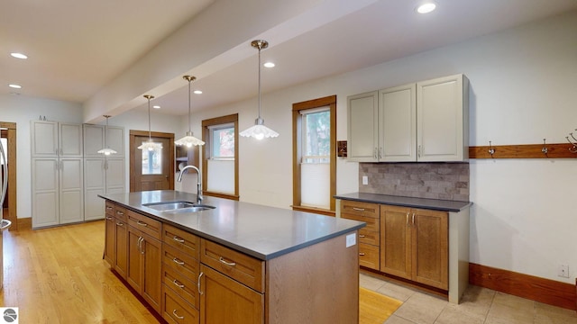 kitchen featuring sink, light wood-type flooring, an island with sink, decorative light fixtures, and decorative backsplash