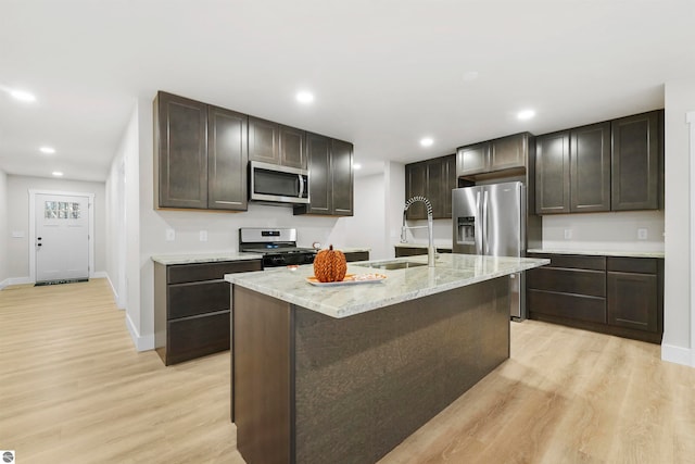 kitchen with dark brown cabinets, stainless steel appliances, a kitchen island with sink, sink, and light hardwood / wood-style floors