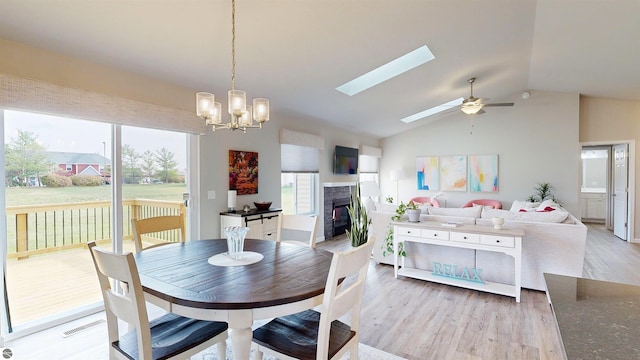 dining space with a tile fireplace, vaulted ceiling with skylight, ceiling fan with notable chandelier, and light wood-type flooring