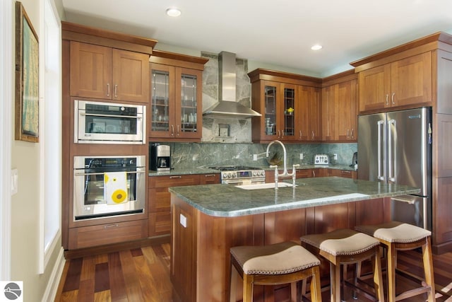 kitchen featuring dark wood-type flooring, wall chimney range hood, sink, an island with sink, and tasteful backsplash