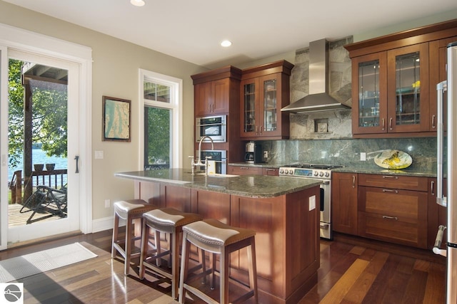 kitchen with dark hardwood / wood-style flooring, wall chimney exhaust hood, stainless steel appliances, sink, and a center island with sink