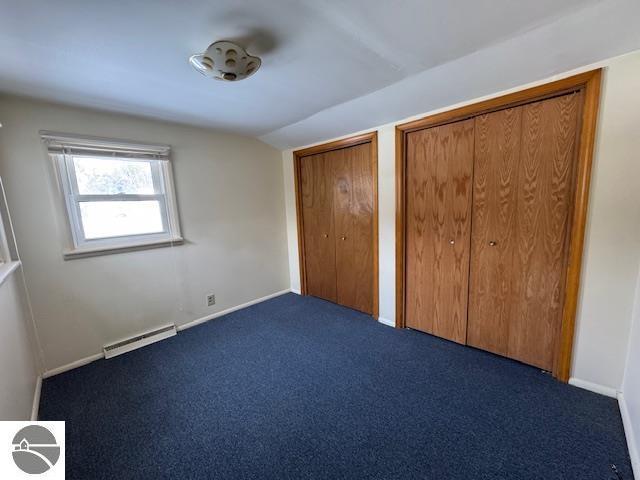 unfurnished bedroom featuring vaulted ceiling, baseboard heating, dark carpet, and multiple closets