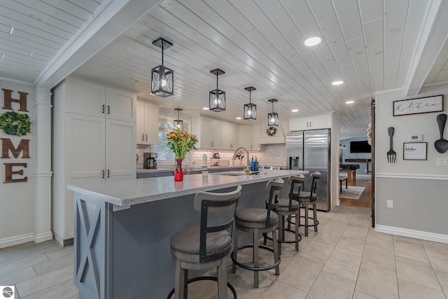 kitchen featuring pendant lighting, stainless steel refrigerator with ice dispenser, white cabinetry, an island with sink, and light stone counters