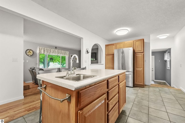 kitchen with light tile patterned flooring, sink, stainless steel fridge, a kitchen island with sink, and a textured ceiling