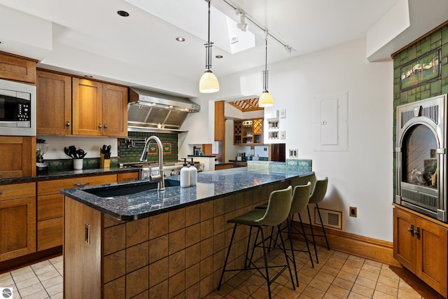 kitchen with stainless steel microwave, ventilation hood, dark stone counters, hanging light fixtures, and a kitchen island with sink