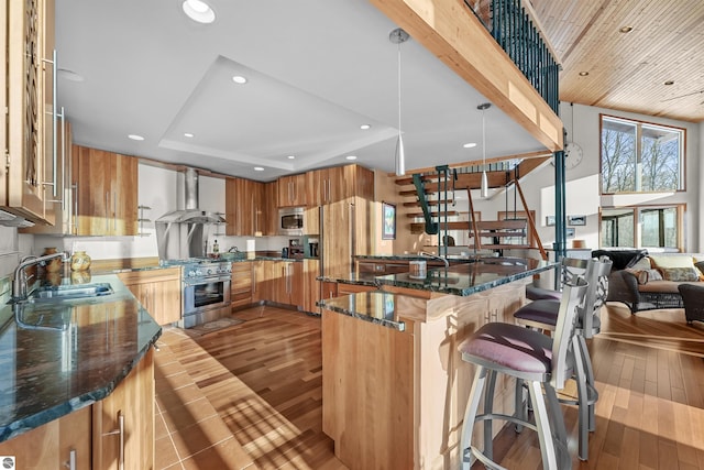 kitchen with wall chimney exhaust hood, sink, built in appliances, hanging light fixtures, and dark stone counters