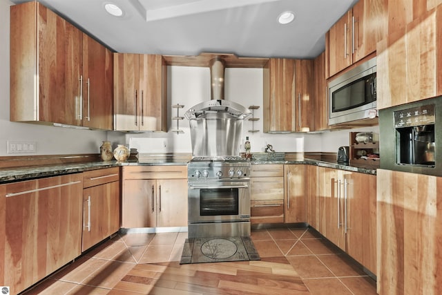 kitchen with stainless steel appliances, dark tile patterned floors, dark stone countertops, and wall chimney exhaust hood