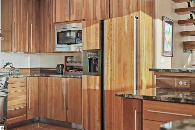 kitchen with stainless steel microwave, dark tile patterned flooring, paneled fridge with ice dispenser, and dark stone counters