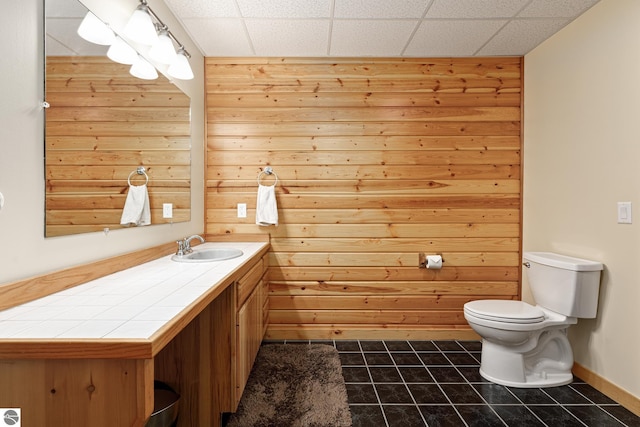 bathroom with a paneled ceiling, tile patterned flooring, wooden walls, vanity, and toilet