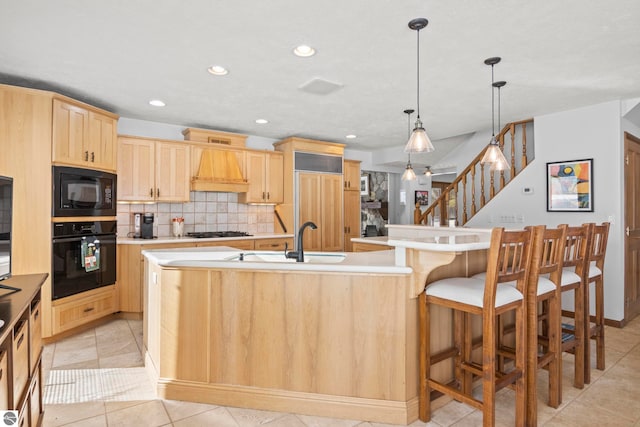 kitchen featuring a center island with sink, light brown cabinets, custom range hood, pendant lighting, and black appliances