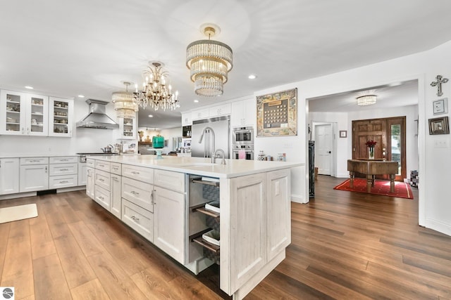 kitchen featuring wall chimney exhaust hood, white cabinetry, decorative light fixtures, appliances with stainless steel finishes, and an island with sink