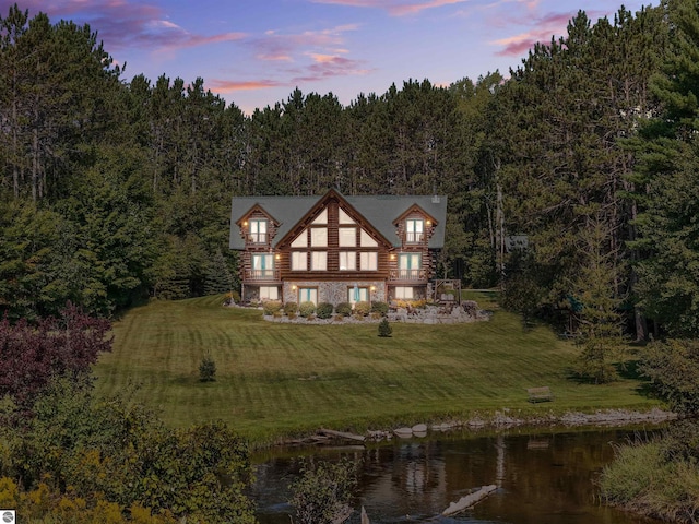 rear view of property featuring stone siding, a water view, a lawn, and a wooded view