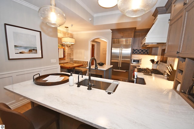 kitchen featuring light stone counters, ornamental molding, stainless steel built in refrigerator, premium range hood, and a sink