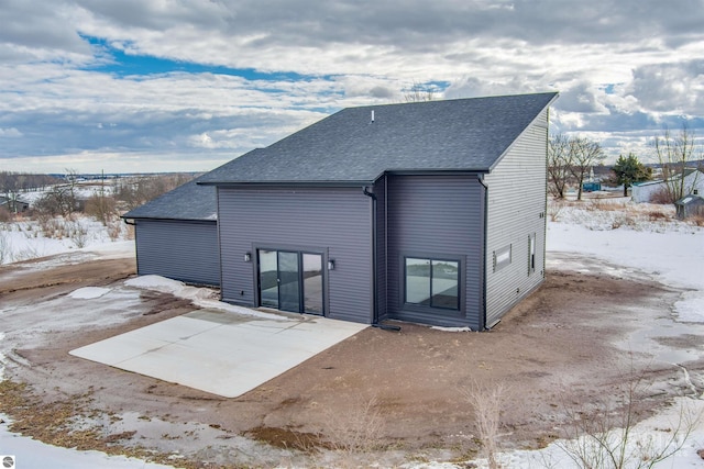 snow covered rear of property with a patio and a shingled roof