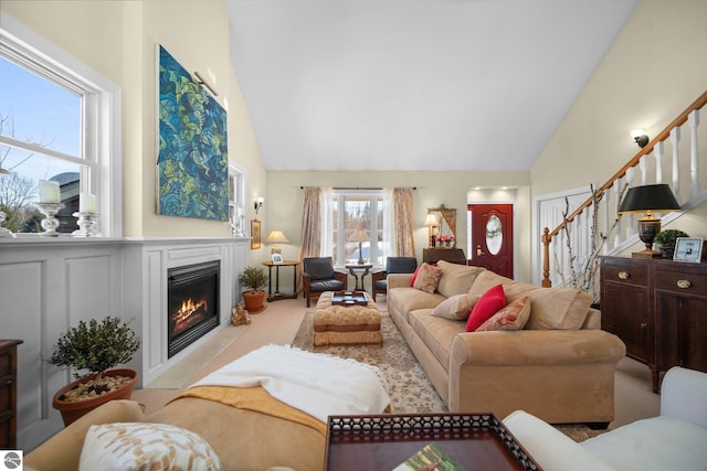 carpeted living area featuring high vaulted ceiling, a glass covered fireplace, and stairs