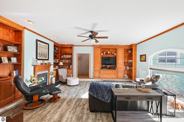 living area with built in shelves, wood finished floors, a ceiling fan, a glass covered fireplace, and crown molding