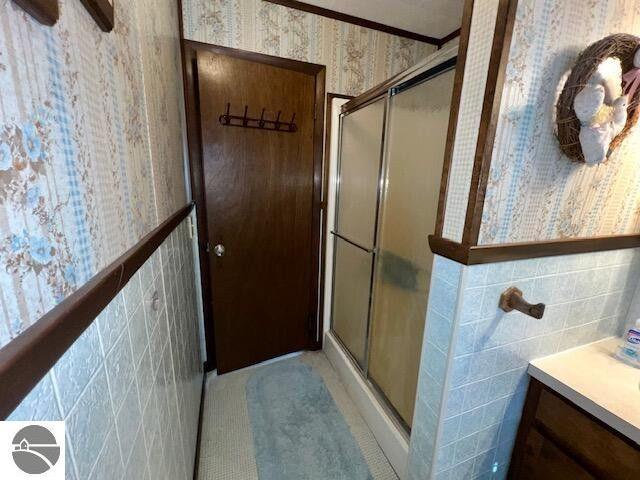 full bath featuring tile walls, a stall shower, wainscoting, vanity, and wallpapered walls