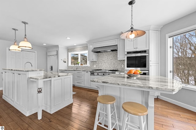 kitchen with a spacious island, appliances with stainless steel finishes, backsplash, range hood, and hardwood / wood-style floors