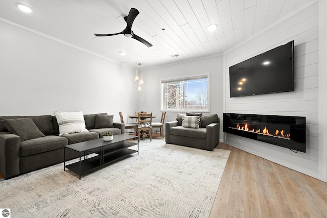 living room with ceiling fan, wooden ceiling, wood finished floors, a glass covered fireplace, and crown molding