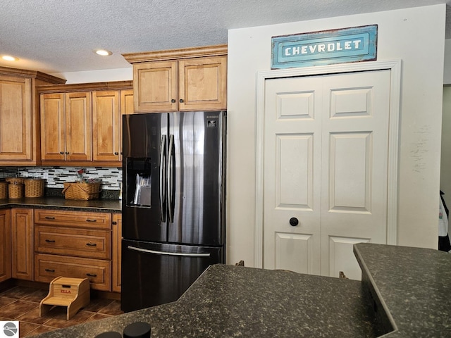 kitchen featuring backsplash, brown cabinetry, dark stone counters, dark tile patterned flooring, and black fridge