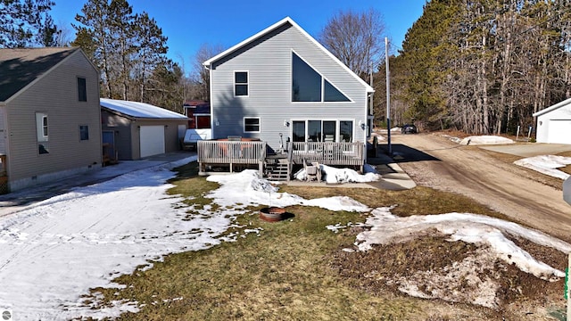 snow covered house with a wooden deck, a detached garage, and an outbuilding