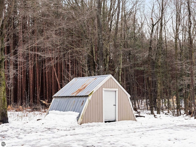 snow covered structure featuring an outbuilding, a forest view, and a storage shed