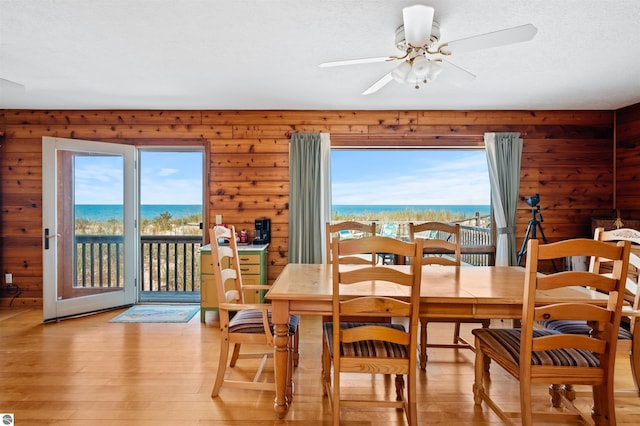 dining area with wooden walls, a ceiling fan, light wood-style flooring, a water view, and a textured ceiling