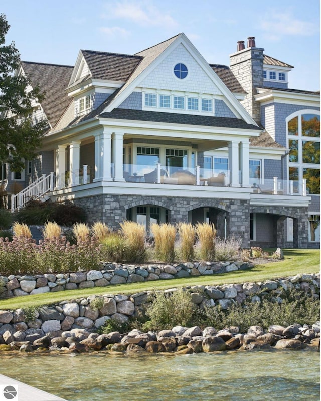 back of property with a shingled roof, a balcony, stone siding, and a chimney