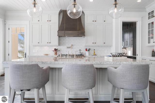 kitchen featuring glass insert cabinets, crown molding, a center island with sink, white cabinetry, and wall chimney exhaust hood